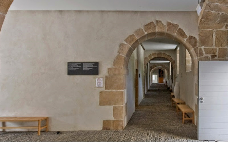 corredor de um edifício antigo com paredes brancas, arcos adornados e piso coberto em pedras rústicas
