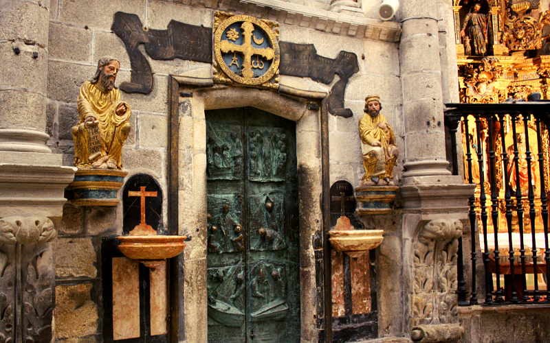 porta de igreja antiga ladeada por eculturas de dois homens e duas pias de água benta