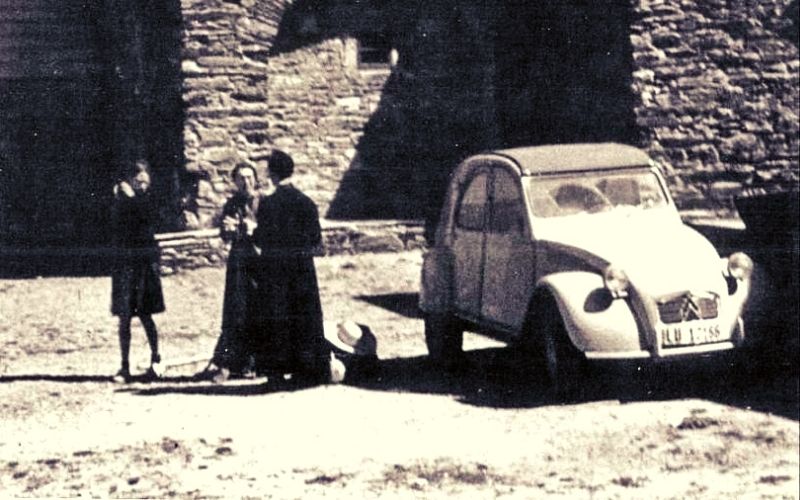 imagem antiga em branco e preto com três pessoas conversando ao lado de um carro e em frente a uma igreja