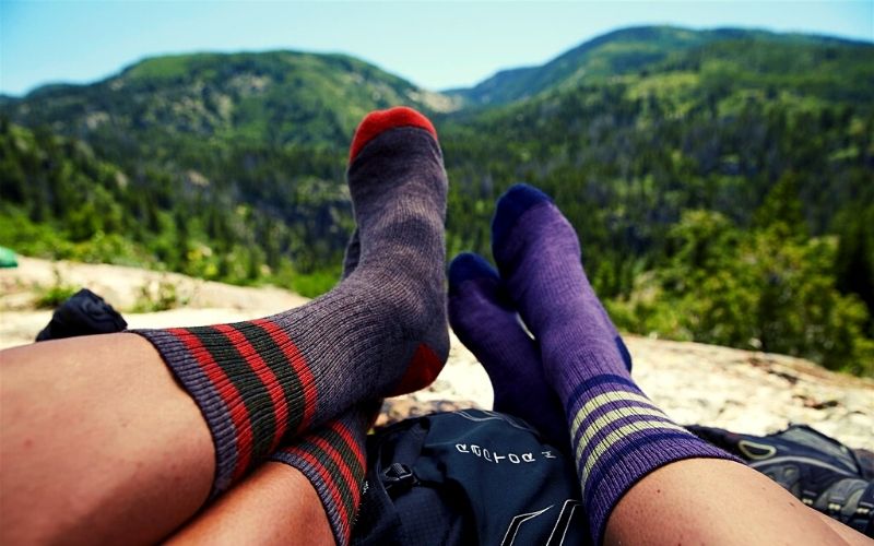 duas pessoas descansando os pés com mkeias sobre mochilas e paisagem natural ao fundo