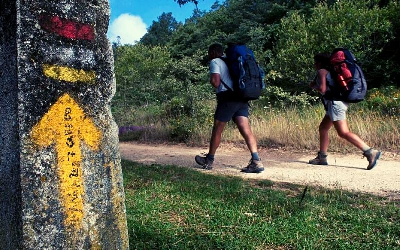 duas pessoas caminhando com mochila nas costas seguindo a seta amarela, sinalização símbolo do Caminho de Santiago,