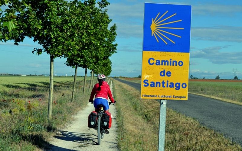 mulher de bicicleta, de costas, numa trilha senalizada como sendo o Caminho de Santiago