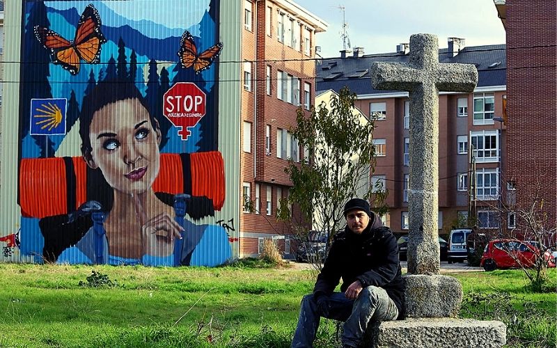 homem sentado sob uma cruz diante do mural de uma mulher