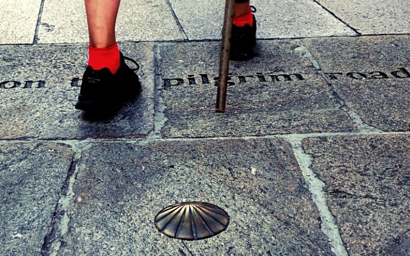 pés calçados de tênis preto e meia vermelha caminhando com a ajuda de um cajado, em calçada de pedra onde há uma concha de metal e esculpido em baixo relevo a palavra pilgrim