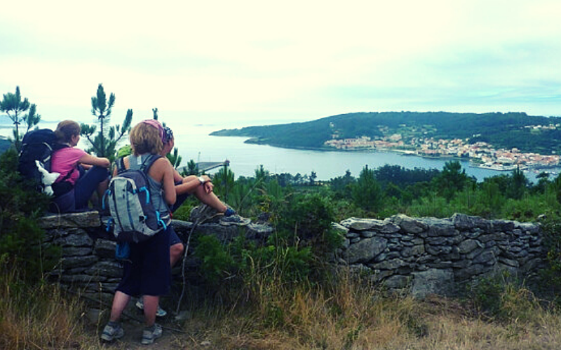 peregrinos sentados sobre uma mureta de pedras apreciando a paisagem com o mar ao fundo