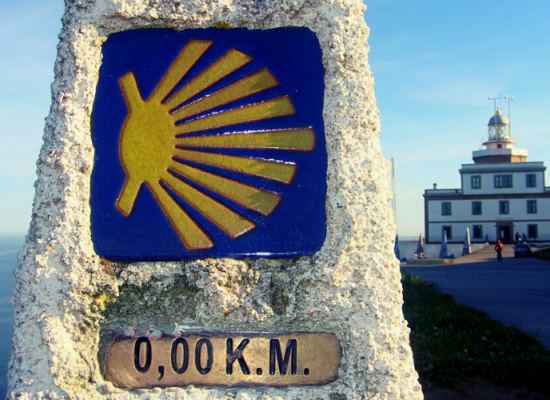 marco zero feito em pedra sinalizado com uma concha amarela sob um fundo azul e ao fundo o farol de finisterre