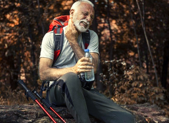 homem grisalho sentado com mochila nas costas garrafa de agua em uma das mãos e bastões de caminhada ao lado