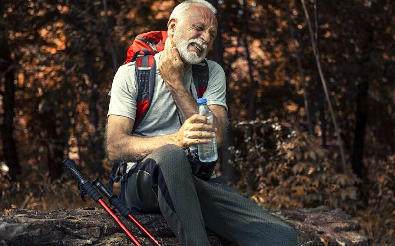 homem grisalho sentado com mochila nas costas garrafa de agua em uma das mãos e bastões de caminhada ao lado